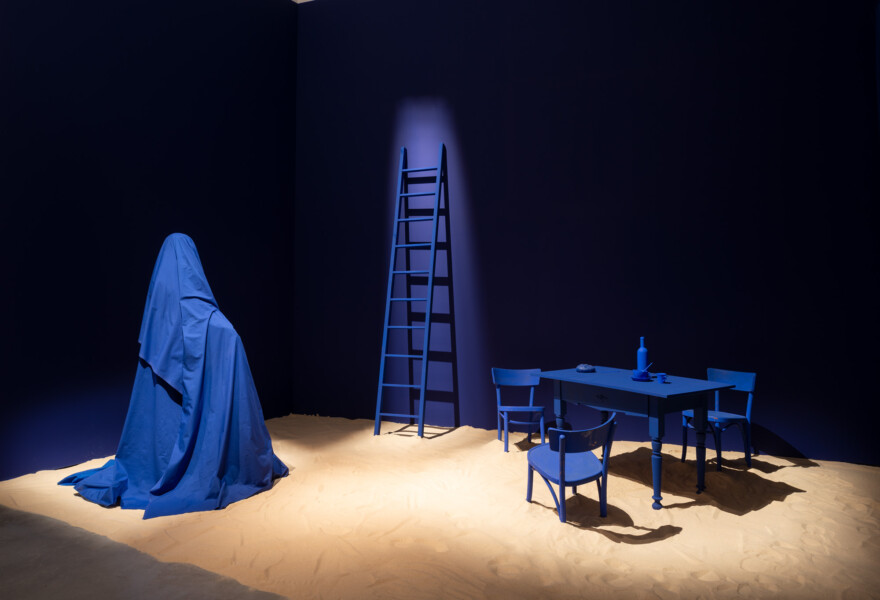 instalación de arte de Eielson con una mesa y cuatro sillas, en la mesa una botella, un plato y una taza. Al lado una escalera y un pedazo de cuero cubriendo un objeto. Todos los elementos son azules índigo. El suelo está cubierto de arena y las paredes están pintadas de negro