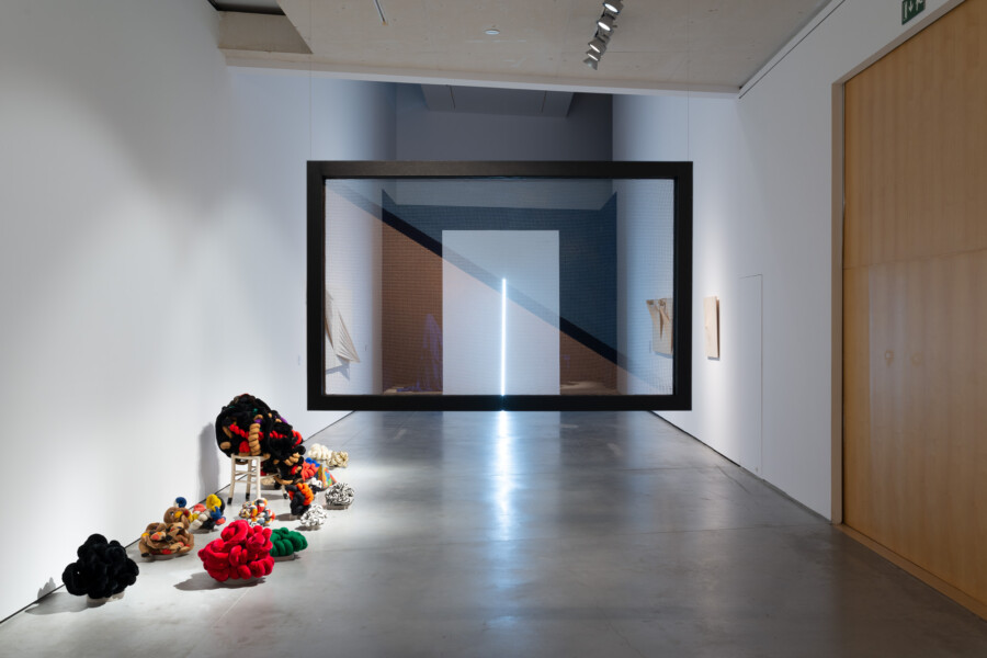 una sala de museo con esculturas de tela colorida anundada a la izquierda y en el centro de la sala un gran rectángulo negro por el que se puede ver a través