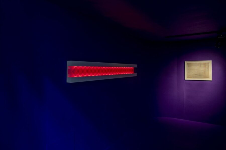 un rectángulo de luz led roja que refleja los latidos del corazón de una artista. Colgado en un muro color violeta. A su lado un boceta de esta obra de arte