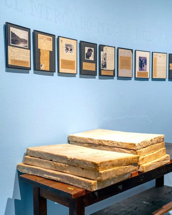 Nury González, El mercado negro del jabón, 1999. Siete barras de 17 kg de jabón sobre mesón de madera, diez fotografías y documentos enmarcados, baúl de mimbre, bolso de cuero, bolsa de tela con objetos en su interior y texto a punto cruz dibujado sobre el muro. Colección Il Posto. Foto cortesía